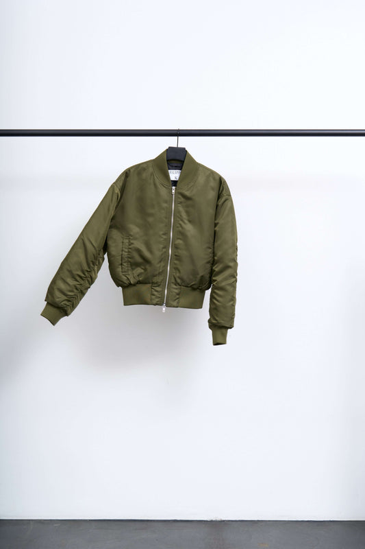 NYLON ZIP JACKET in olive green, lightweight bomber with a clean silhouette, hanging on a rack.